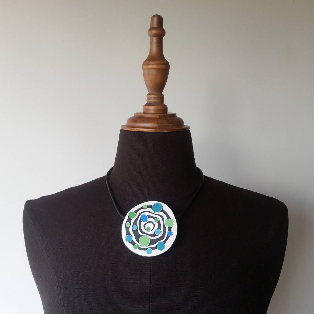 Large handmade pendant with organically-shaped concentric black and white circles, and irregular dots of bright blue and green. It is approximately 6.6cm in diameter and hangs on a black adjustable cord.